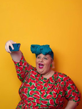 Artı deniz mavisi bir başlık takan ve karpuz elbiseli bir kadın canlı turuncu bir arka plan önünde akıllı telefonuyla selfie çekerken gülümsüyor..