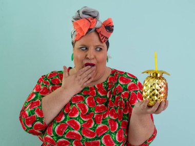 Karpuz desenli bir elbise giymiş tuhaf bir kadın ve elinde altın ananas şeklindeki bir kupayla kırmızı güneş gözlüklerinin üzerinden neşeyle bakan bir kupa tutuyor..