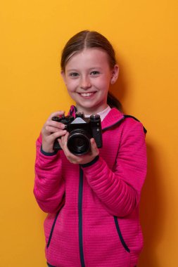 Pembe kapüşonlu şirin bir kız elinde eski model bir kamera tutarken parlak turuncu stüdyo arka planına karşı duruyor.. 