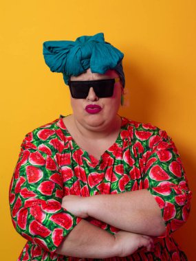 Kendine güvenen, karpuz desenli elbise ve deniz mavisi kafa örtüsü takan, koyu renk güneş gözlüğü takan, turuncu bir arka plana karşı ciddi bir ifadeyle poz veren iri yarı bir kadın..