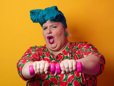 Artı renkli karpuz desenli elbise ve deniz mavisi başlıklı kadın turuncu bir arka plana karşı komik ve konsantre bir yüz ifadesiyle pembe halterleri kaldırıyor..