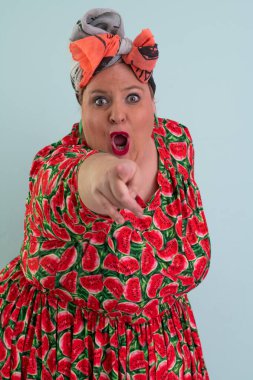 Plus size woman in a colorful watermelon themed dress and orange headwrap posing with unimpressed facial expression and hands on hips against pastel blue background.  clipart