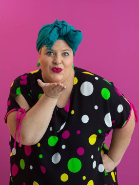 Puantiyeli elbise ve deniz mavisi başlıklı neşeli bir yüz ifadesiyle canlı pembe arka planda öpüşen neşeli, iri yarı bir kadın.. 