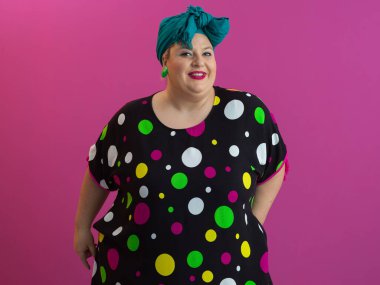 Neşeli, puantiyeli elbise ve deniz mavisi başlıklı kadın canlı pembe bir arka plana karşı büyük bir gülümsemeyle poz veriyor, pozitifliği, vücut güvenini ve neşeli tavırları temsil ediyor.. 