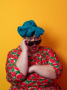 Kendine güvenen, karpuz desenli elbise ve deniz mavisi kafa örtüsü takan, koyu renk güneş gözlüğü takan, turuncu bir arka plana karşı ciddi bir ifadeyle poz veren iri yarı bir kadın..