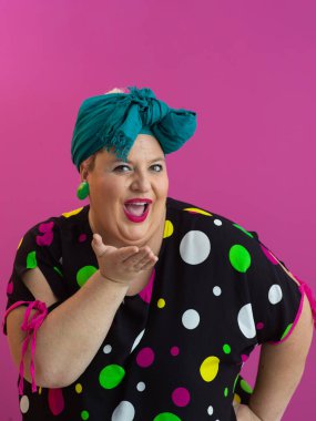 Puantiyeli elbise ve deniz mavisi başlıklı neşeli bir yüz ifadesiyle canlı pembe arka planda öpüşen neşeli, iri yarı bir kadın.. 