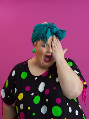 Parlak rujlu, mavi bantlı, tek gözünü puantiyeli elbisesi olan, neşeli pembe bir arka planda poz veren, parlak saçlı, iri yarı bir kadın.