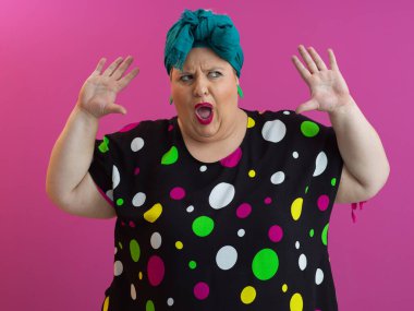 Renkli puantiyeli elbiseler içinde etkileyici, iri yarı bir kadın canlı pembe arka plana karşı komik bir sürprizle el kaldırıyor.. 