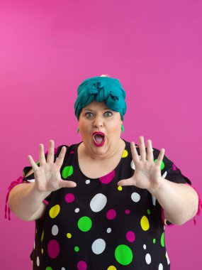 Expressive plus size woman in colorful polka dot dress raising hands in humorous surprise against vibrant pink background.  clipart