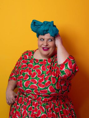 Artı karpuz temalı bir elbise giyen boyunda bir kadın ve kafasına dokunurken neşeli poz veren deniz mavisi başlıklı bir kadın, canlı turuncu bir arka plana karşı..