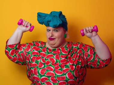 Artı renkli karpuz desenli elbise ve deniz mavisi başlıklı kadın turuncu bir arka plana karşı komik ve konsantre bir yüz ifadesiyle pembe halterleri kaldırıyor..