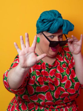 Kendine güvenen, karpuz desenli elbise ve deniz mavisi kafa örtüsü takan, koyu renk güneş gözlüğü takan, turuncu bir arka plana karşı ciddi bir ifadeyle poz veren iri yarı bir kadın..