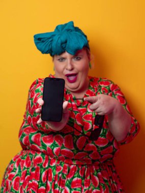 Heyecanlı ve cüsseli bir kadın deniz mavisi bir başlık takıyor ve karpuz elbisesini tutuyor ve renkli bir stüdyo ortamında telefonuna neşeli bir ifadeyle işaret ediyor..