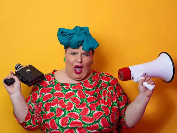 Elinde megafon ve klasik bir kamera olan karpuz elbiseli, etkileyici, iri yarı bir kadın parlak turuncu bir arka plan önünde enerjik bir şekilde bağırıyor..