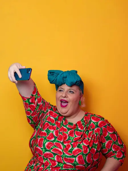 Artı deniz mavisi bir başlık takan ve karpuz elbiseli bir kadın canlı turuncu bir arka plan önünde akıllı telefonuyla selfie çekerken gülümsüyor..