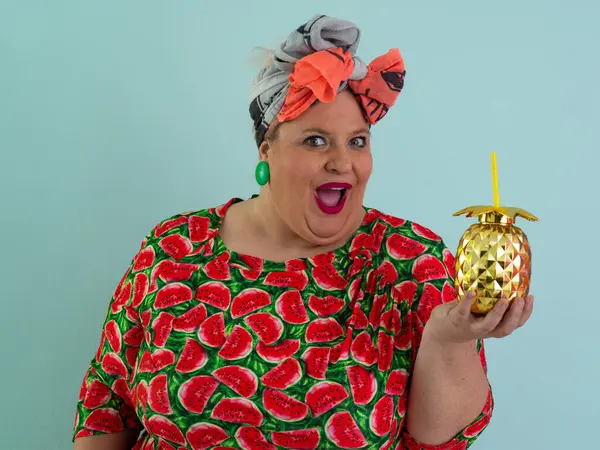 Karpuz desenli bir elbise giymiş tuhaf bir kadın ve elinde altın ananas şeklindeki bir kupayla kırmızı güneş gözlüklerinin üzerinden neşeyle bakan bir kupa tutuyor..