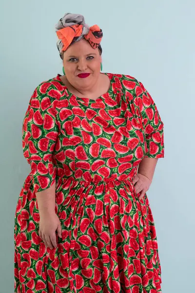 Ayrıca renkli karpuz temalı elbisesi ve turuncu başlıklı, etkilenmemiş yüz ifadesi ve pastel mavisi arka planda kalçaları olan bir kadın.. 