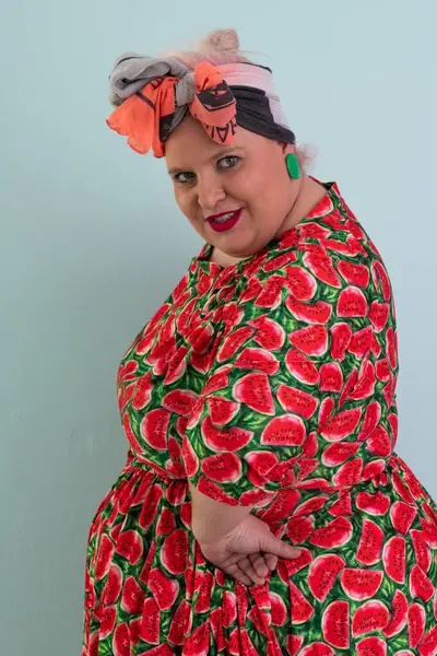 Ayrıca renkli karpuz temalı elbisesi ve turuncu başlıklı, etkilenmemiş yüz ifadesi ve pastel mavisi arka planda kalçaları olan bir kadın.. 