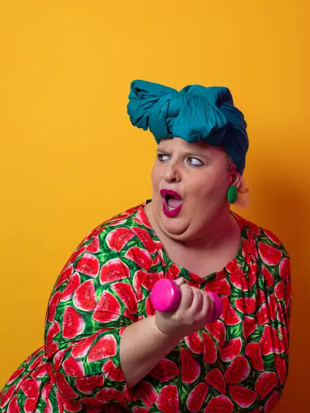 Artı karpuz elbisesi ve pembe halterli yeşil başlıklı kadın. Alnını silerken turuncu arka planda egzersiz yaptıktan sonra yorgunluk ve rahatlama belirtileri gösteriyor..