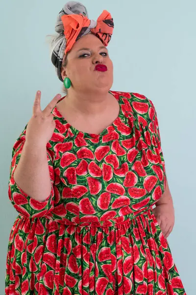 Ayrıca renkli karpuz temalı elbisesi ve turuncu başlıklı, etkilenmemiş yüz ifadesi ve pastel mavisi arka planda kalçaları olan bir kadın.. 