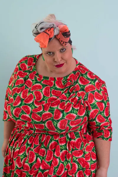 Ayrıca renkli karpuz temalı elbisesi ve turuncu başlıklı, etkilenmemiş yüz ifadesi ve pastel mavisi arka planda kalçaları olan bir kadın.. 