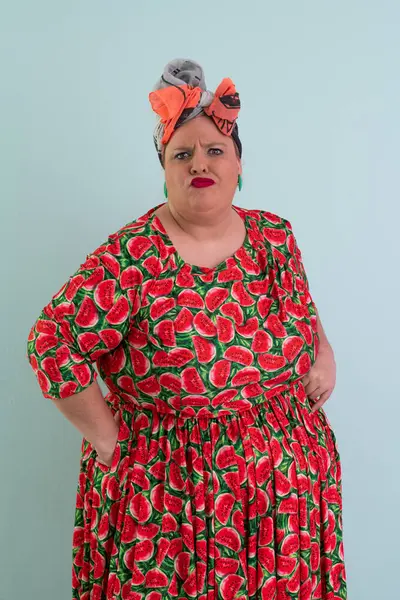 Ayrıca renkli karpuz temalı elbisesi ve turuncu başlıklı, etkilenmemiş yüz ifadesi ve pastel mavisi arka planda kalçaları olan bir kadın.. 