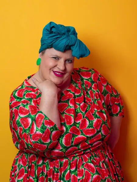 Artı karpuz temalı bir elbise giyen boyunda bir kadın ve kafasına dokunurken neşeli poz veren deniz mavisi başlıklı bir kadın, canlı turuncu bir arka plana karşı..