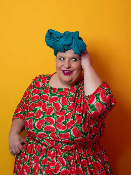 Artı karpuz temalı bir elbise giyen boyunda bir kadın ve kafasına dokunurken neşeli poz veren deniz mavisi başlıklı bir kadın, canlı turuncu bir arka plana karşı..