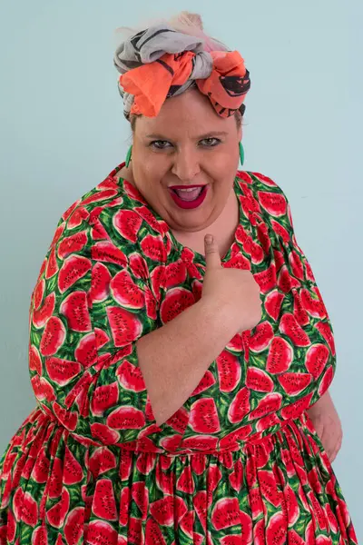 Ayrıca renkli karpuz temalı elbisesi ve turuncu başlıklı, etkilenmemiş yüz ifadesi ve pastel mavisi arka planda kalçaları olan bir kadın.. 