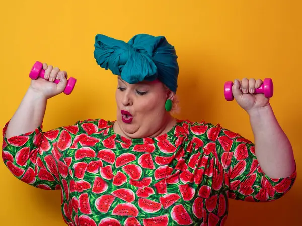 Artı renkli karpuz desenli elbise ve deniz mavisi başlıklı kadın turuncu bir arka plana karşı komik ve konsantre bir yüz ifadesiyle pembe halterleri kaldırıyor..