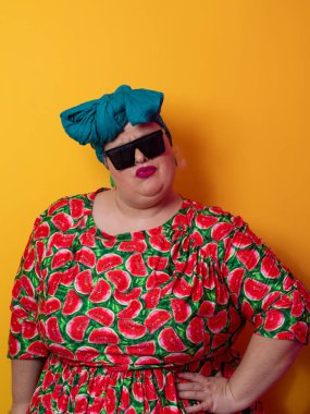 Kendine güvenen, karpuz desenli elbise ve deniz mavisi kafa örtüsü takan, koyu renk güneş gözlüğü takan, turuncu bir arka plana karşı ciddi bir ifadeyle poz veren iri yarı bir kadın..