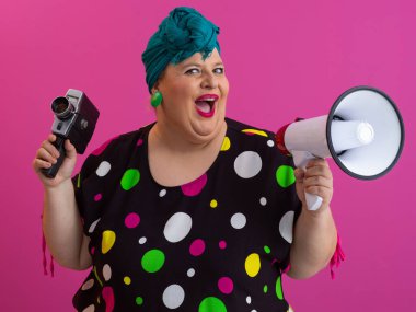 Canlı pembe bir arkaplan önünde elinde eski model bir kamerayla megafona bağıran komik, iri yarı bir kadın.. 