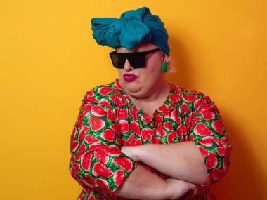 Kendine güvenen, karpuz desenli elbise ve deniz mavisi kafa örtüsü takan, koyu renk güneş gözlüğü takan, turuncu bir arka plana karşı ciddi bir ifadeyle poz veren iri yarı bir kadın..
