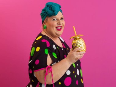 Kendine güveni tam ve eskimiş bir kadın elinde altın bir ananas bardağı tutarken parlak pembe bir arka plana karşı tavır takınıyor.. 