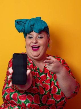 Heyecanlı ve cüsseli bir kadın deniz mavisi bir başlık takıyor ve karpuz elbisesini tutuyor ve renkli bir stüdyo ortamında telefonuna neşeli bir ifadeyle işaret ediyor..