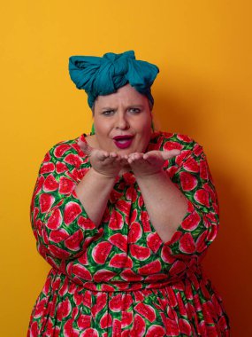 Artı karpuz temalı bir elbise giyen boyunda bir kadın ve kafasına dokunurken neşeli poz veren deniz mavisi başlıklı bir kadın, canlı turuncu bir arka plana karşı..