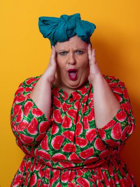 Artı karpuz temalı bir elbise giyen boyunda bir kadın ve kafasına dokunurken neşeli poz veren deniz mavisi başlıklı bir kadın, canlı turuncu bir arka plana karşı..