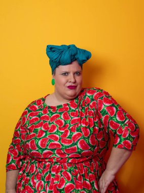 Artı karpuz temalı bir elbise giyen boyunda bir kadın ve kafasına dokunurken neşeli poz veren deniz mavisi başlıklı bir kadın, canlı turuncu bir arka plana karşı..