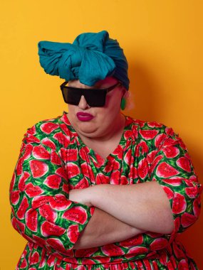 Kendine güvenen, karpuz desenli elbise ve deniz mavisi kafa örtüsü takan, koyu renk güneş gözlüğü takan, turuncu bir arka plana karşı ciddi bir ifadeyle poz veren iri yarı bir kadın..