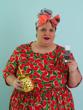 Karpuz desenli bir elbise giymiş tuhaf bir kadın ve elinde altın ananas şeklindeki bir kupayla kırmızı güneş gözlüklerinin üzerinden neşeyle bakan bir kupa tutuyor.. 