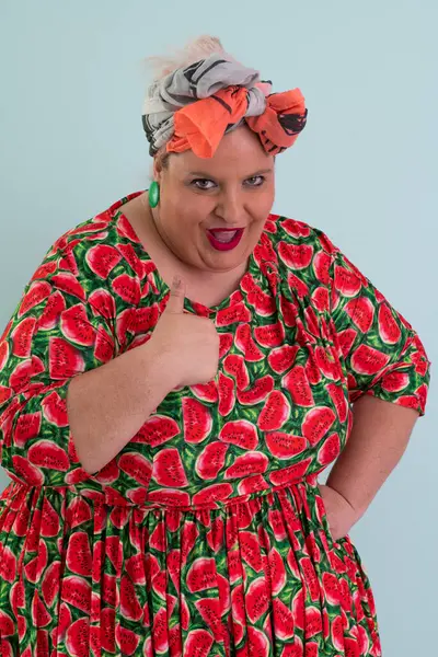 Ayrıca renkli karpuz temalı elbisesi ve turuncu başlıklı, etkilenmemiş yüz ifadesi ve pastel mavisi arka planda kalçaları olan bir kadın.. 