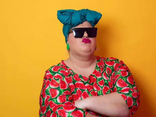 Kendine güvenen, karpuz desenli elbise ve deniz mavisi kafa örtüsü takan, koyu renk güneş gözlüğü takan, turuncu bir arka plana karşı ciddi bir ifadeyle poz veren iri yarı bir kadın..