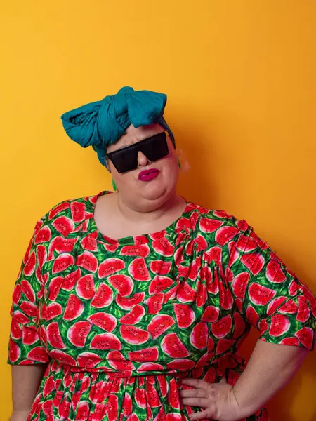 Kendine güvenen, karpuz desenli elbise ve deniz mavisi kafa örtüsü takan, koyu renk güneş gözlüğü takan, turuncu bir arka plana karşı ciddi bir ifadeyle poz veren iri yarı bir kadın..