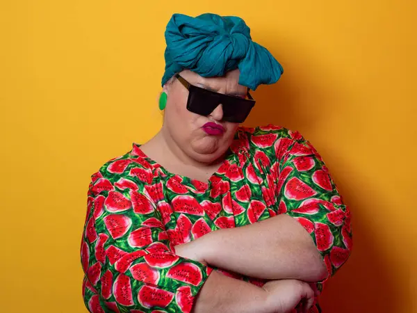 Kendine güvenen, karpuz desenli elbise ve deniz mavisi kafa örtüsü takan, koyu renk güneş gözlüğü takan, turuncu bir arka plana karşı ciddi bir ifadeyle poz veren iri yarı bir kadın..