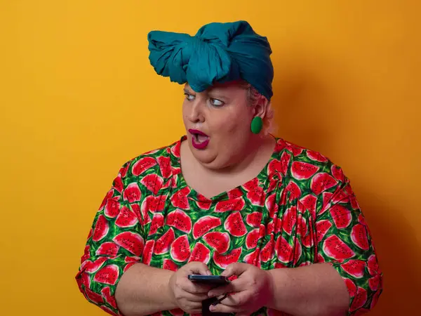 Artı deniz mavisi bir başlık ve karpuz elbise giyen bir kadın canlı turuncu bir arka planda akıllı telefonuna şaşırmış bir ifadeyle bakıyor..