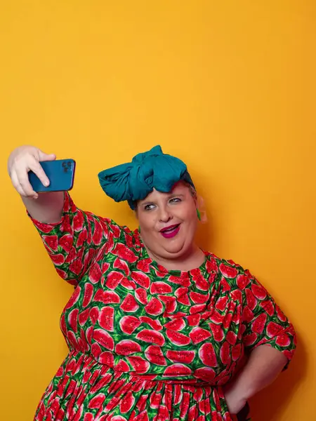 Artı deniz mavisi bir başlık takan ve karpuz elbiseli bir kadın canlı turuncu bir arka plan önünde akıllı telefonuyla selfie çekerken gülümsüyor..