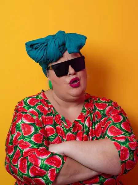 Kendine güvenen, karpuz desenli elbise ve deniz mavisi kafa örtüsü takan, koyu renk güneş gözlüğü takan, turuncu bir arka plana karşı ciddi bir ifadeyle poz veren iri yarı bir kadın..