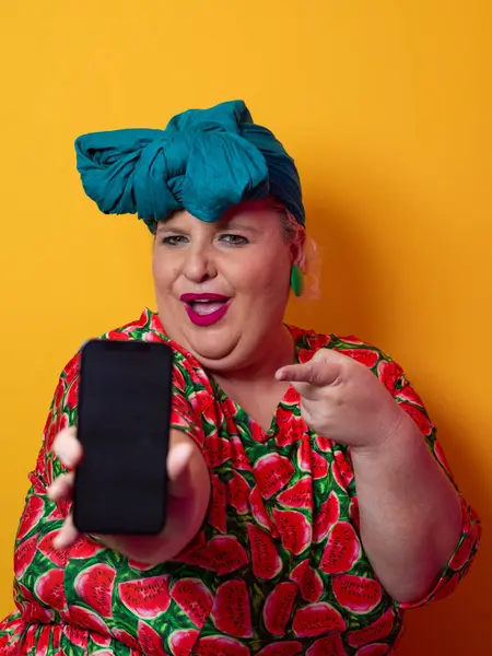 Heyecanlı ve cüsseli bir kadın deniz mavisi bir başlık takıyor ve karpuz elbisesini tutuyor ve renkli bir stüdyo ortamında telefonuna neşeli bir ifadeyle işaret ediyor..