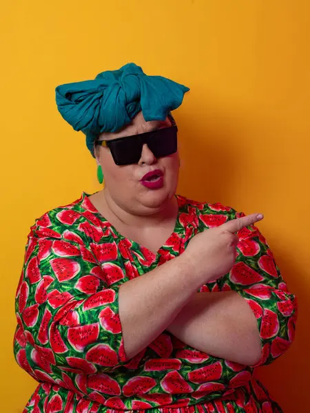 Kendine güvenen, karpuz desenli elbise ve deniz mavisi kafa örtüsü takan, koyu renk güneş gözlüğü takan, turuncu bir arka plana karşı ciddi bir ifadeyle poz veren iri yarı bir kadın..
