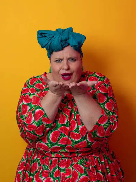 Artı karpuz temalı bir elbise giyen boyunda bir kadın ve kafasına dokunurken neşeli poz veren deniz mavisi başlıklı bir kadın, canlı turuncu bir arka plana karşı..